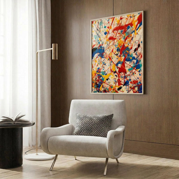 May include: Abstract expressionist artwork with vibrant red, blue, yellow, and orange paint splatters and drips on cream background, displayed in thin frame above modern beige armchair with geometric pillow. Room features wood paneled wall, brass floor lamp, and side table with open book.