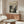May include: A framed Jackson Pollock exhibition poster hanging above a mid-century modern wooden credenza in a minimalist living room. The vintage-style poster features "POLLOCK" in bold black letters with "CHAOS AND CREATIVITY 1912-1956" and displays an abstract black and white drip painting. The room has neutral beige walls, a white textured chair, glass-top coffee table, and modern lighting fixtures creating warm shadows on the polished floor.