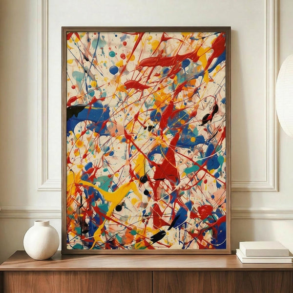 May include: Abstract expressionist artwork with energetic paint splatters, drips and gestural brushstrokes in vibrant red, blue, yellow and black against cream background. Framed in thin brown frame, displayed on wooden sideboard next to white ceramic vase and stacked books in elegant room with white paneled walls.