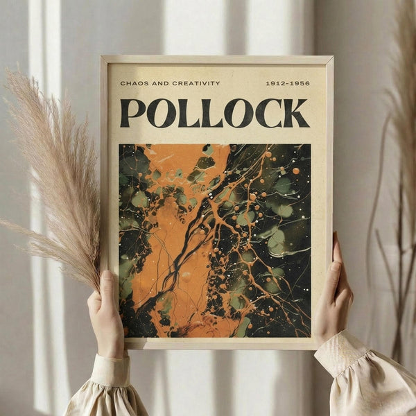 May include: Person in cream ruffle-sleeve top holding framed abstract art poster featuring dynamic orange and dark green paint splatters and drips. Dried pampas grass visible on left side. Light wooden frame against neutral background with soft natural lighting.