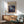 May include: Framed poster featuring Jackson Pollock's abstract expressionist artwork with swirling blue, purple, and white paint drips in cosmic patterns. Displayed above mid-century modern wooden credenza with white ceramic lamp, next to sage green upholstered armchair with wooden frame. Warm natural lighting creates shadows on white wall in contemporary living room setting.