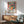 May include: Framed poster with colorful still life artwork featuring flowers in yellow vase, fruits, and pottery in Matisse-inspired style with bold reds, greens, and blues. Displayed above wooden sideboard with white ceramic lamp in modern living room with sage green upholstered chair and round coffee table. Bright natural lighting creates geometric shadows on white wall.