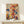 May include: Abstract expressionist artwork with energetic splatters, drips and gestural marks in vibrant red, blue, yellow and black paint over cream background, displayed in thin natural wood frame on light wooden console table beside small white wrapped gift with evergreen sprig against white wall.