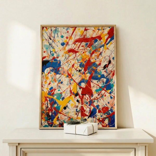 May include: Abstract expressionist artwork with energetic splatters, drips and gestural marks in vibrant red, blue, yellow and black paint over cream background, displayed in thin natural wood frame on light wooden console table beside small white wrapped gift with evergreen sprig against white wall.