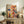May include: Vibrant abstract expressionist artwork with chaotic splatters and drips in red, blue, yellow, and black on cream background, displayed in thin wooden frame on rustic wood sideboard. Room setting shows brown leather chair, fern plant, ceramic vase, and stacked books creating warm, cozy interior atmosphere.