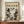 May include: Framed vintage-style poster with "POLLOCK" and "CHAOS AND CREATIVITY 1912-1956" text above abstract black and white paint splatter artwork. Brown wooden frame displayed on light wood sideboard next to white ceramic vase and books against cream walls with white wainscoting.