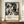 May include: Black and white abstract expressionist artwork poster in thin wooden frame, displayed on walnut sideboard next to cream ceramic vase and stacked books. The poster features chaotic paint splatters and drips in Jackson Pollock's signature style, with exhibition text below reading "Jackson Pollock January 10-20, 1956 8th Avenue, New York City A Journey Through Chaos." Set against cream walls with white wainscoting in elegant interior.