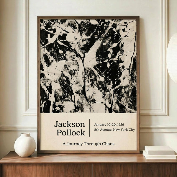 May include: Black and white abstract expressionist artwork poster in thin wooden frame, displayed on walnut sideboard next to cream ceramic vase and stacked books. The poster features chaotic paint splatters and drips in Jackson Pollock's signature style, with exhibition text below reading "Jackson Pollock January 10-20, 1956 8th Avenue, New York City A Journey Through Chaos." Set against cream walls with white wainscoting in elegant interior.