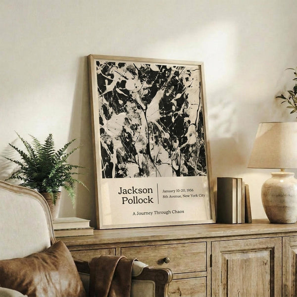 May include: Black and white abstract Jackson Pollock exhibition poster in light wood frame, displayed on rustic wooden credenza alongside fern plant, books, and ceramic vase in neutral-toned living room with beige chair and brown throw pillows