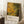 May include: A framed abstract art poster with organic yellow and green circular patterns resembling cells or bubbles, displayed on a white textured rug against white paneled walls. The poster shows Jackson Pollock exhibition details at bottom in cream section. A wooden side table with white coffee cup and saucer sits nearby, with beige fabric visible on right edge.