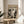 May include: Framed poster featuring abstract black and white artwork with dynamic paint splatters and flowing forms, displayed on a light wood round side table next to a beige upholstered chair. Natural sunlight streams across the minimalist room with white walls and a green plant visible in the corner.