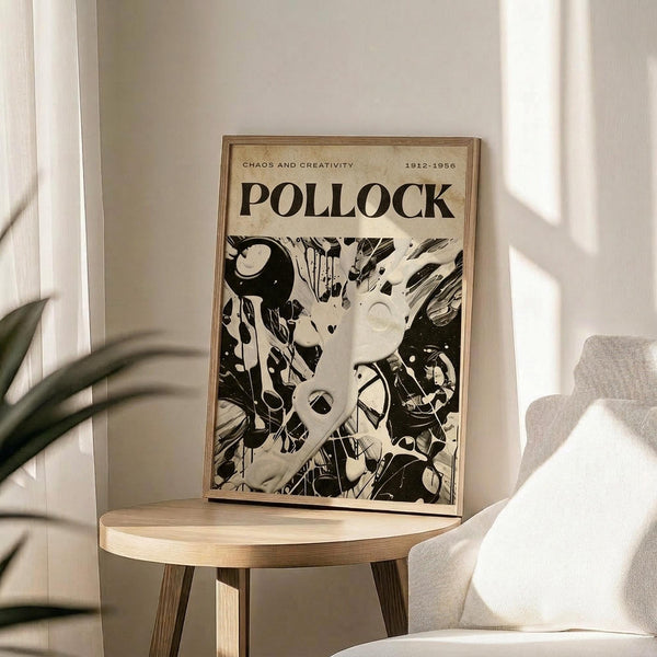 May include: Framed poster featuring abstract black and white artwork with dynamic paint splatters and flowing forms, displayed on a light wood round side table next to a beige upholstered chair. Natural sunlight streams across the minimalist room with white walls and a green plant visible in the corner.