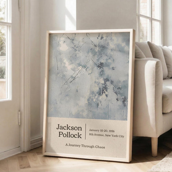 May include: Framed abstract art poster with pale blue and gray brushstrokes leaning against white wall next to beige sofa. The artwork features organic splatters and linear marks in muted tones. Exhibition-style text appears below the abstract image on cream background within light wood frame on hardwood floor.