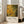 May include: Framed poster print featuring abstract artwork with flowing yellow and green organic shapes, circular bubbles, and dark branching lines resembling tree branches or neural networks. The piece has an earthy, natural color palette transitioning from golden yellow to deep green. Below the artwork is white text reading "Jackson Pollock" with exhibition details and "A Journey Through Chaos." The poster leans against a white wall next to a beige upholstered sofa in a bright, minima