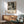 May include: Black and white abstract expressionist artwork in wooden frame hanging above mid-century modern wooden sideboard with white ceramic lamp. Sage green upholstered armchair with wooden arms sits nearby on hardwood floor. Natural sunlight creates dramatic shadows across white wall. The framed poster features chaotic paint splatters and drips in monochrome palette with exhibition text below the artwork.