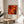 May include: Framed art print featuring Jackson Pollock's abstract expressionist style with vibrant orange, red, and yellow paint splatters and drips against dark areas. The poster shows exhibition details and sits on a light wood round side table next to a beige upholstered chair. Soft natural light filters through white curtains, casting gentle shadows. A plant's green leaves are partially visible in the corner, creating a warm, modern living room setting.