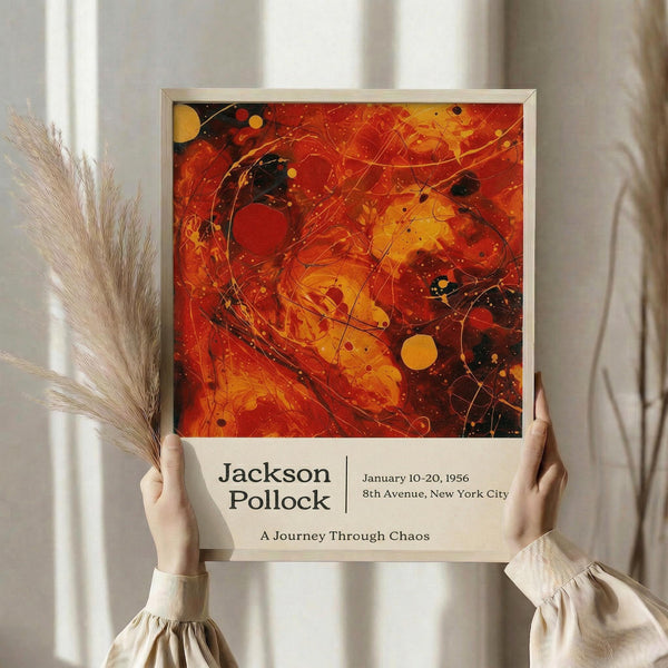 May include: Person in beige ruffled sleeves holding framed abstract art poster featuring vibrant red, orange, and yellow swirling paint with black accents and circular elements. White matted frame against neutral curtained background with dried pampas grass visible on left side.