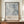 May include: Framed abstract art poster with pale blue and gray watercolor-style brushstrokes and black linear elements, displayed on wooden sideboard next to cream ceramic vase and stacked books. Text reads "Jackson Pollock January 10-20, 1956 8th Avenue, New York City A Journey Through Chaos" in brown lettering below artwork. Neutral beige wall background.