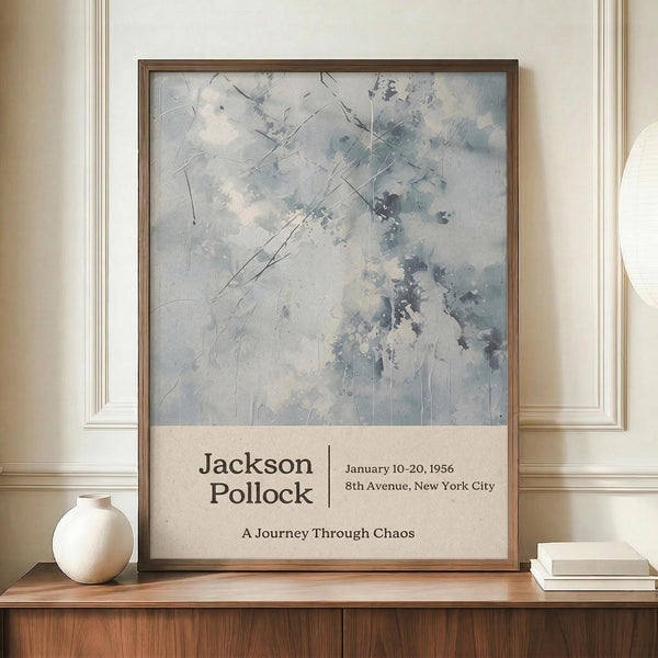 May include: Framed abstract art poster with pale blue and gray watercolor-style brushstrokes and black linear elements, displayed on wooden sideboard next to cream ceramic vase and stacked books. Text reads "Jackson Pollock January 10-20, 1956 8th Avenue, New York City A Journey Through Chaos" in brown lettering below artwork. Neutral beige wall background.
