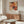 May include: Abstract expressionist art poster with vibrant orange, red, black and white paint splatters and drips displayed above a mid-century modern wooden credenza with sliding doors. White upholstered chair and glass-top coffee table visible in minimalist living room with beige walls and natural lighting.
