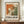 May include: Framed exhibition poster featuring Jackson Pollock-style abstract artwork with orange, green, and white paint splatters and drips. Bold black text reads "POLLOCK" and "EMBRACE THE CHAOTIC REALM OF CREATIVITY" with exhibition dates. Warm wooden frame sits on mid-century credenza beside white ceramic vase and stacked books against cream walls with white trim molding.