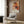 May include: Abstract art poster featuring orange, red, and black paint splatters with white geometric lines, displayed in thin frame on wood-paneled wall above beige upholstered armchair with geometric patterned pillow. Modern living room setting with brass floor lamp and side table.