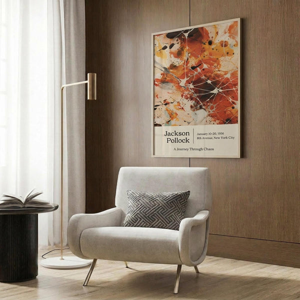 May include: Abstract art poster featuring orange, red, and black paint splatters with white geometric lines, displayed in thin frame on wood-paneled wall above beige upholstered armchair with geometric patterned pillow. Modern living room setting with brass floor lamp and side table.