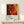 May include: Jackson Pollock exhibition poster mockup in natural wood frame, featuring abstract artwork with vibrant red, orange, yellow and black paint splatters and circular forms. Poster displays exhibition details and "A Journey Through Chaos" text below artwork. Frame leans against white wall on light wooden shelf next to small white wrapped gift with twine and evergreen sprig.