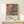 May include: Framed abstract art poster with energetic paint splatters in orange, blue, yellow and black colors, displayed on white wooden console table against cream wall. Small white wrapped gift with evergreen sprig sits nearby. Artwork shows dynamic drip painting style with flowing lines and vibrant color combinations in thin gold frame.