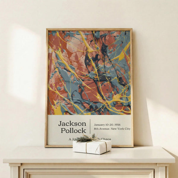 May include: Framed abstract art poster with energetic paint splatters in orange, blue, yellow and black colors, displayed on white wooden console table against cream wall. Small white wrapped gift with evergreen sprig sits nearby. Artwork shows dynamic drip painting style with flowing lines and vibrant color combinations in thin gold frame.