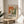 May include: Framed exhibition poster featuring abstract orange, green and white paint splatters in Jackson Pollock's drip painting style. Text reads "POLLOCK EMBRACE THE CHAOTIC REALM OF CREATIVITY" with exhibition dates May 18, 2030 - August 14, 2030. Natural wood frame sits on round wooden side table next to beige upholstered chair with plant leaves visible, bright sunlight streaming across white walls.