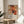 May include: Abstract expressionist artwork poster in natural wood frame displaying orange, red, black and white paint splatters and geometric line overlays. Poster sits on round wooden side table next to beige upholstered chair with throw pillow. Plant leaves visible in corner, white walls with natural window light creating shadows across the minimalist modern interior setting.