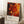 May include: Framed art poster with abstract paint splatter design in vibrant reds, oranges, and yellows with dark accents and circular bubble patterns, displayed on white fluffy rug against white wall. Features exhibition-style text at bottom. Coffee cup on wooden block visible nearby, creating cozy room mockup scene.