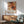 May include: Abstract art poster with orange, red, and black paint splatters and white geometric line overlays, displayed in thin wood frame above mid-century modern credenza with white ceramic lamp. Mint green upholstered chair with wooden arms sits nearby on wooden floors with natural sunlight casting shadows across white wall.