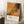 May include: Framed abstract art poster with orange and black paint splatters in Jackson Pollock's drip painting style, leaning against white wall next to beige armchair. White coffee cup on wooden side table. Cream-colored frame with white text panel showing exhibition details. Soft natural lighting creates window shadows on wall. White textured rug underneath.