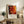 May include: Framed abstract art poster with vibrant red, orange, and black splattered paint design in warm wood frame, displayed on rustic wooden sideboard alongside fern plant, ceramic vase, and leather armchair in cozy neutral-toned living room setting with natural lighting