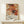 May include: Abstract expressionist art poster with vibrant orange, red, and black paint splatters and drips on white background, displayed in thin wooden frame on light wood console table. White wrapped gift with twine and pine sprig sits in front. Cream colored wall backdrop with soft natural lighting.