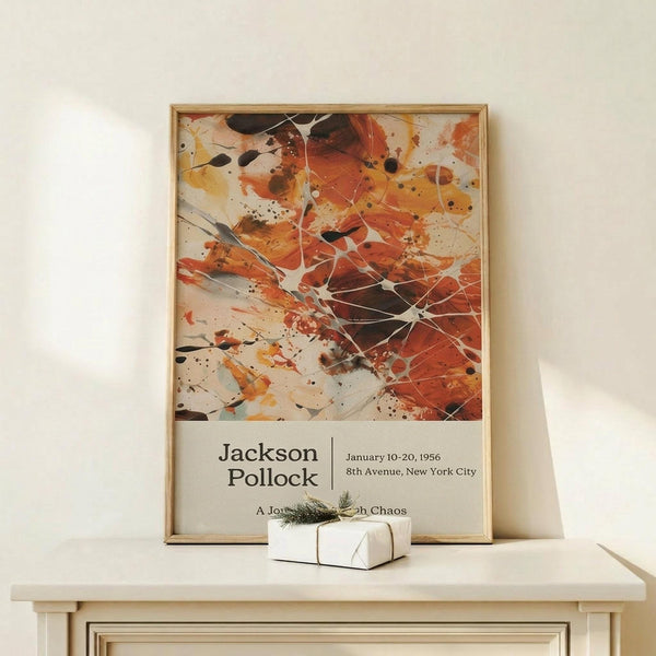 May include: Abstract expressionist art poster with vibrant orange, red, and black paint splatters and drips on white background, displayed in thin wooden frame on light wood console table. White wrapped gift with twine and pine sprig sits in front. Cream colored wall backdrop with soft natural lighting.