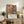 May include: Framed Jackson Pollock exhibition poster with abstract paint splatters in orange, blue, and yellow colors, displayed on rustic wooden sideboard alongside fern plant, ceramic vase, and stacked books in warm-lit living room with beige chair and neutral walls