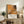 May include: Framed Jackson Pollock exhibition poster with orange and brown abstract splatter artwork displayed on rustic wooden console table alongside fern plant, books, and ceramic vase in neutral-toned living room setting with cream upholstered chair visible in foreground.