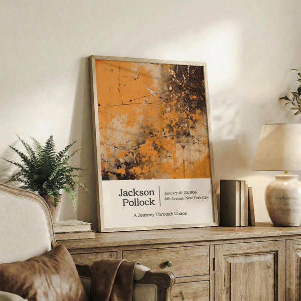 May include: Framed Jackson Pollock exhibition poster with orange and brown abstract splatter artwork displayed on rustic wooden console table alongside fern plant, books, and ceramic vase in neutral-toned living room setting with cream upholstered chair visible in foreground.