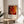 May include: Jackson Pollock exhibition poster featuring abstract artwork with vibrant red, orange, and yellow paint splatters and circular forms against dark background. Poster displays in thin wooden frame on round wooden side table next to beige upholstered chair. Soft natural lighting creates window shadows on neutral wall. Green plant leaves visible in corner of minimalist interior setting.