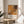 May include: Jackson Pollock exhibition poster with abstract orange and brown paint splatters displayed in thin wooden frame on round wooden side table next to beige upholstered chair. Soft natural lighting creates window shadows on neutral wall. Green plant leaves visible in corner. Poster features artwork reproduction with text below including artist name, exhibition dates, and venue information.