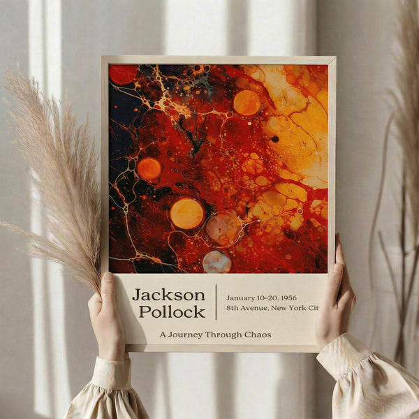 May include: Person holding framed art poster featuring abstract painting with swirling red, orange, and black colors with bubble-like circles throughout. Text below reads "Jackson Pollock" with exhibition details and "A Journey Through Chaos". Light wooden frame against neutral background with dried pampas grass visible to left side.