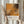 May include: Person in cream sweater holding framed abstract art poster with orange and black paint splatters in Pollock's signature drip style. White frame against soft neutral background with dried pampas grass. Exhibition-style layout with artist name, dates, and "A Journey Through Chaos" text below artwork.