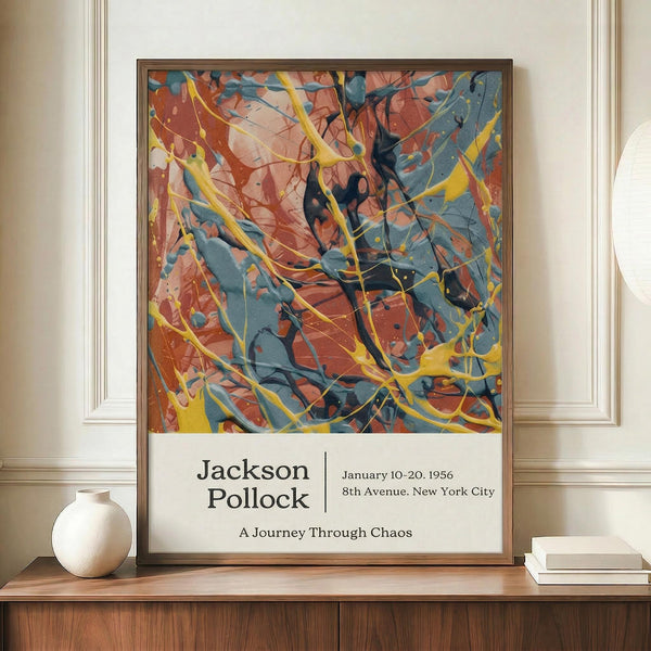 May include: Framed abstract expressionist poster with energetic splashes and drips in vibrant blues, oranges, yellows, and black paint against white background. Exhibition-style text below reads "Jackson Pollock" with dates and location details. Frame sits on wooden credenza beside white ceramic vase and stacked books in neutral-toned room with white paneled walls.