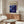 May include: Blue and purple abstract swirled artwork poster with "POLLOCK" text and "1912-1956" dates, displayed in light wood frame above mid-century walnut credenza. Modern living room setting with cream sculptural chair, glass oval coffee table, and white spherical table lamp. Minimalist Scandinavian-style interior with neutral walls and natural lighting.