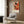 May include: Jackson Pollock exhibition poster with abstract splattered paint in orange, red, black and yellow tones, displayed above a beige upholstered armchair with geometric patterned pillow. Modern living room setting features wood paneled wall, brass floor lamp, dark side table with open book, and light curtains by window.