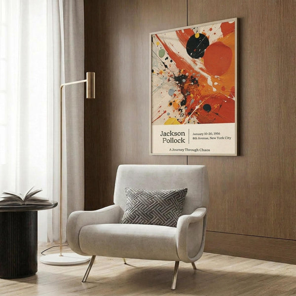 May include: Jackson Pollock exhibition poster with abstract splattered paint in orange, red, black and yellow tones, displayed above a beige upholstered armchair with geometric patterned pillow. Modern living room setting features wood paneled wall, brass floor lamp, dark side table with open book, and light curtains by window.