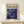 May include: Framed art print featuring swirling cosmic blues and purples with golden accents, displayed on light wood console table next to small wrapped gift with twine bow. Poster shows abstract fluid patterns resembling galaxies or nebulae in deep indigo, violet and cream tones with scattered circular elements. Natural wood frame complements minimal neutral room setting.